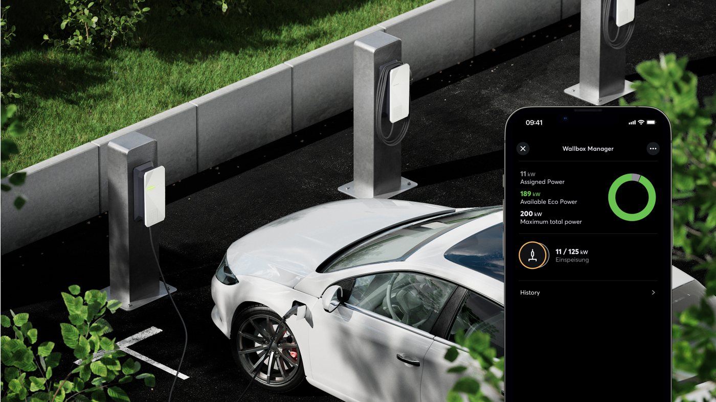 Bílý elektromobil u nabíjecí stanice, vedle mobil s aplikací pro řízení nabíjení